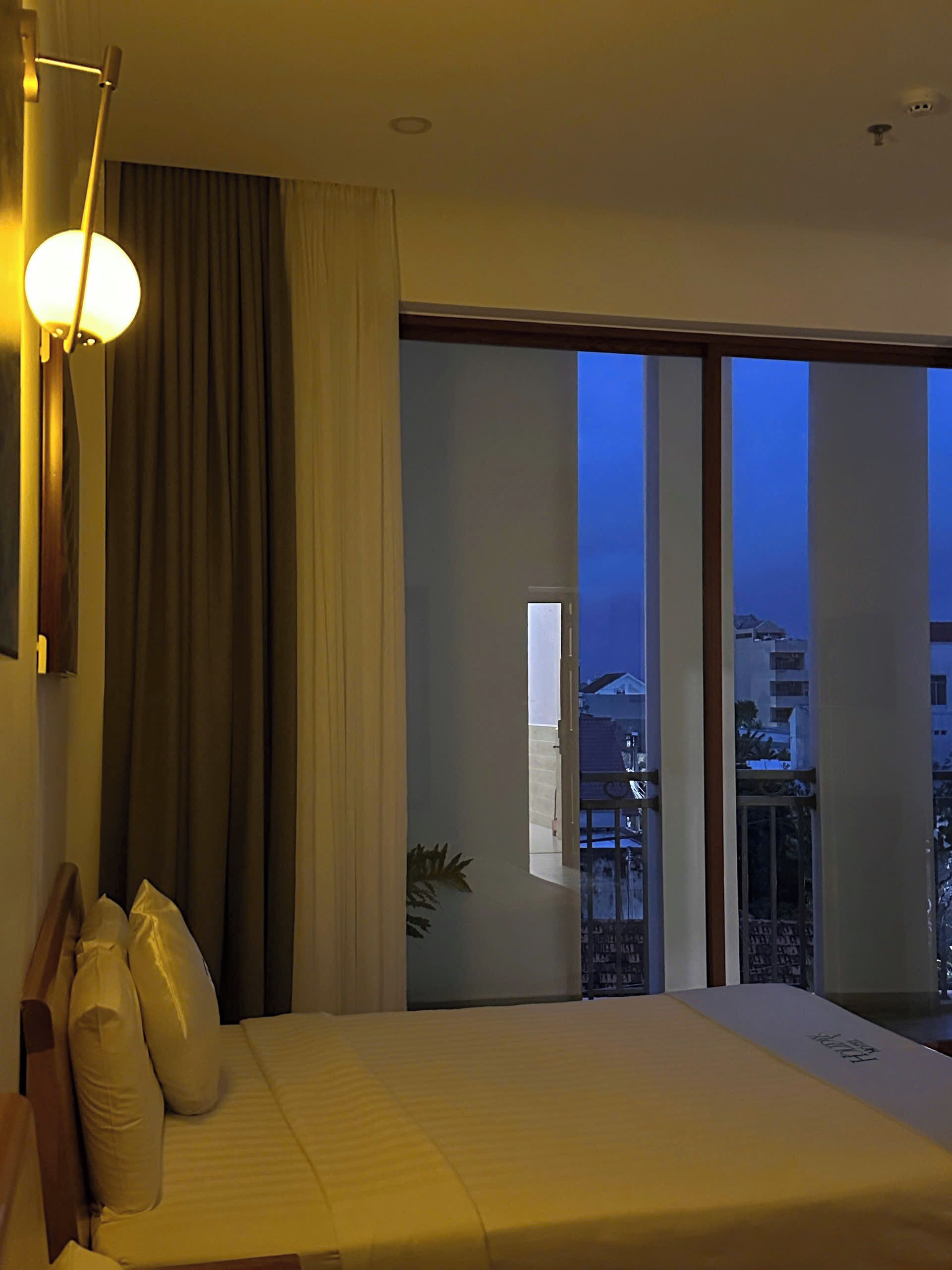 A hotel room with a bed, a large window, and a view of buildings outside.
