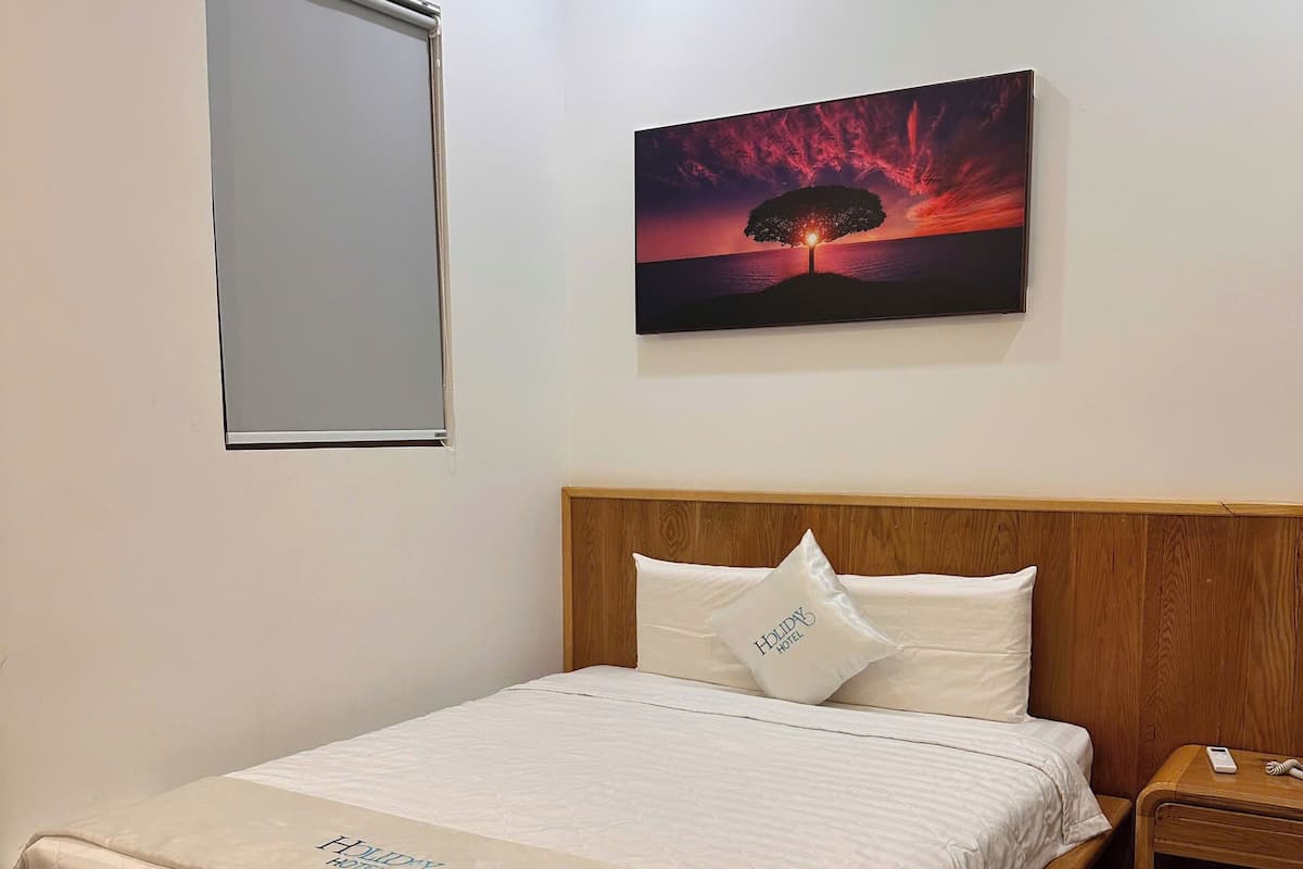 Una cama bien hecha con una cubierta acolchada y una almohada con el nombre del hotel, un cabecero de madera, una mesita de noche con un teléfono y un cuadro de una puesta de sol en la pared.
