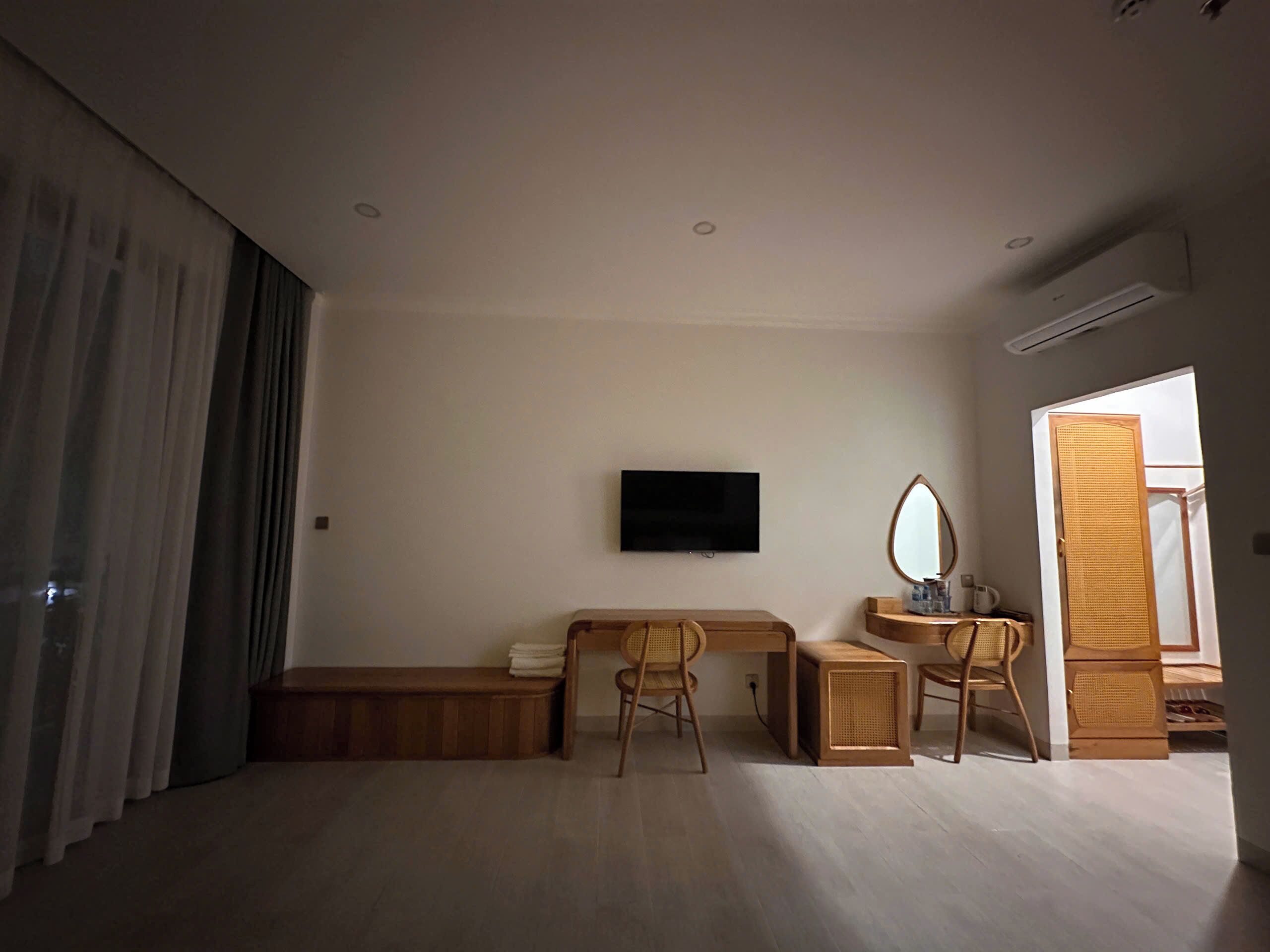 A modern, minimalistic room with a wooden desk, two wicker chairs, a flat-screen TV mounted on the wall, and a wooden wardrobe with a mirror.