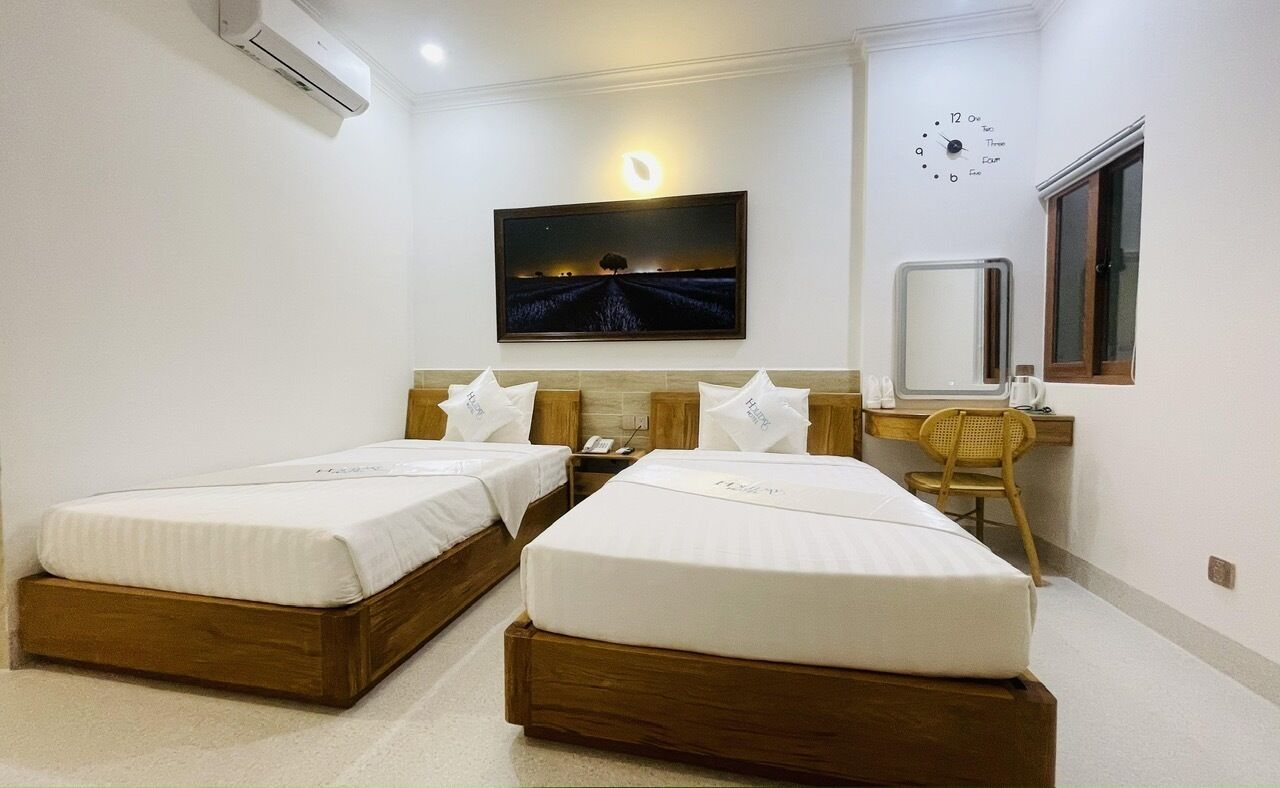 A hotel room with two single beds, a desk with a chair, a wall clock, and a painting of a landscape.