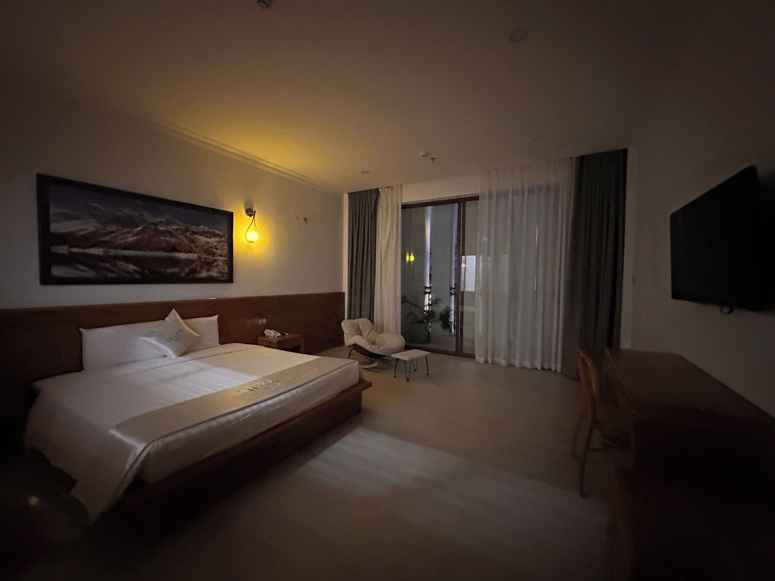 Habitación de hotel con una cama grande, un escritorio, una silla, un televisor y un balcón con cortinas.