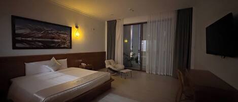 Habitación de hotel con una cama grande, un escritorio, una silla, un televisor y un balcón con cortinas.
