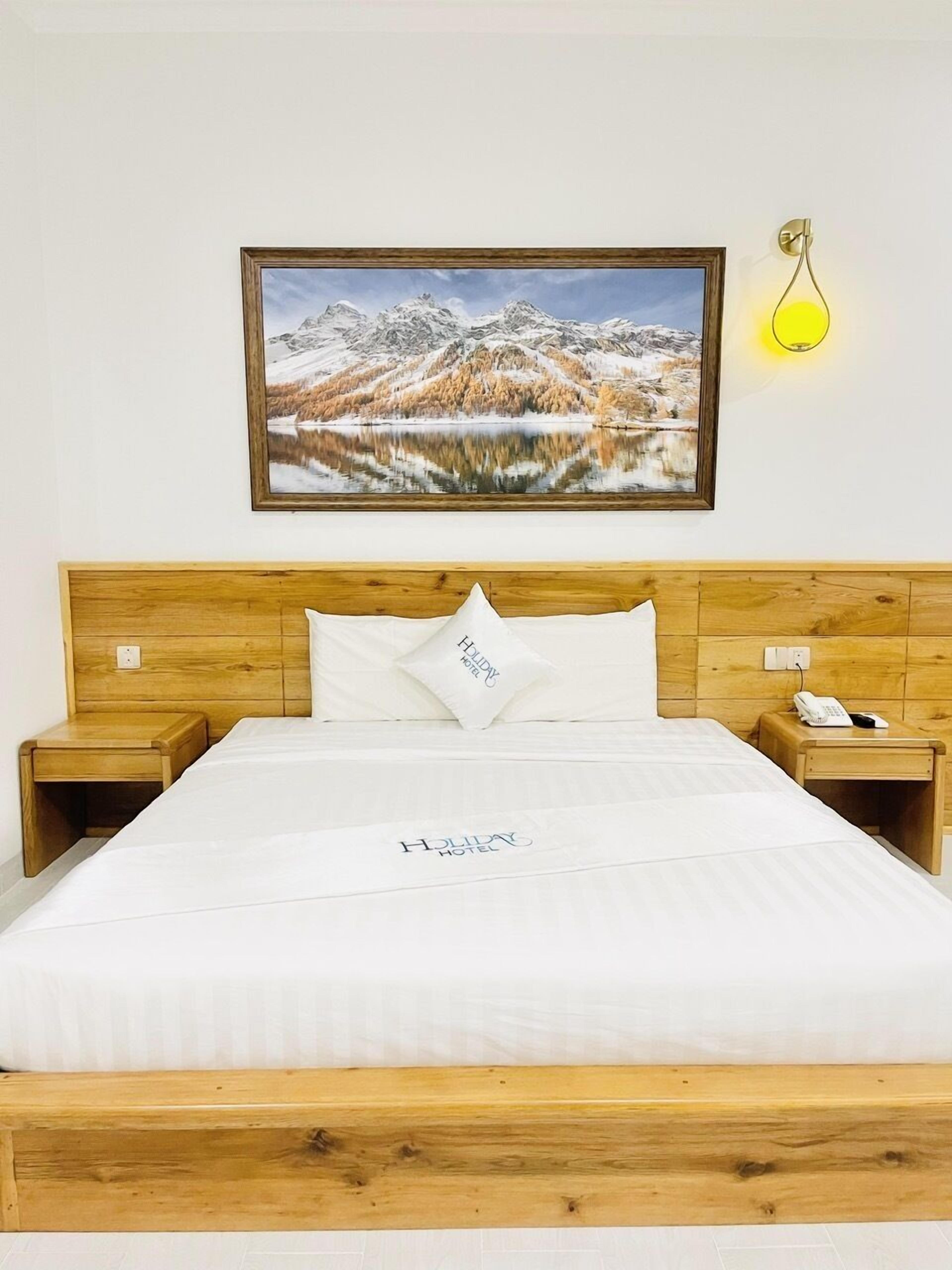 A hotel room with a wooden headboard, a bed with white bedding, a nightstand with a phone, and a painting of a snowy landscape.