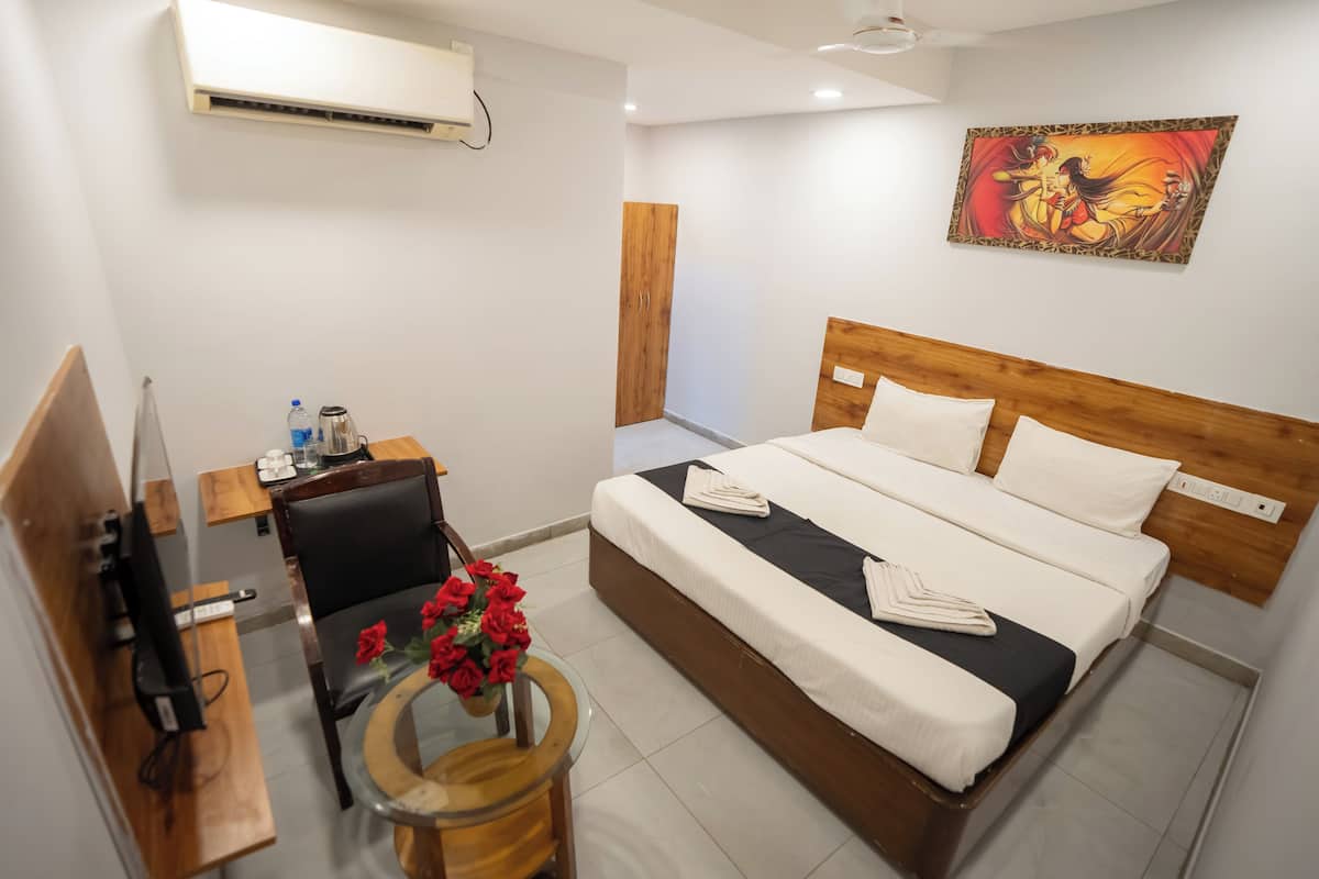 Une chambre d’hôtel comprenant un lit, un bureau avec une chaise, une petite table ornée d’un vase de fleurs rouges et un climatiseur fixé au mur.
