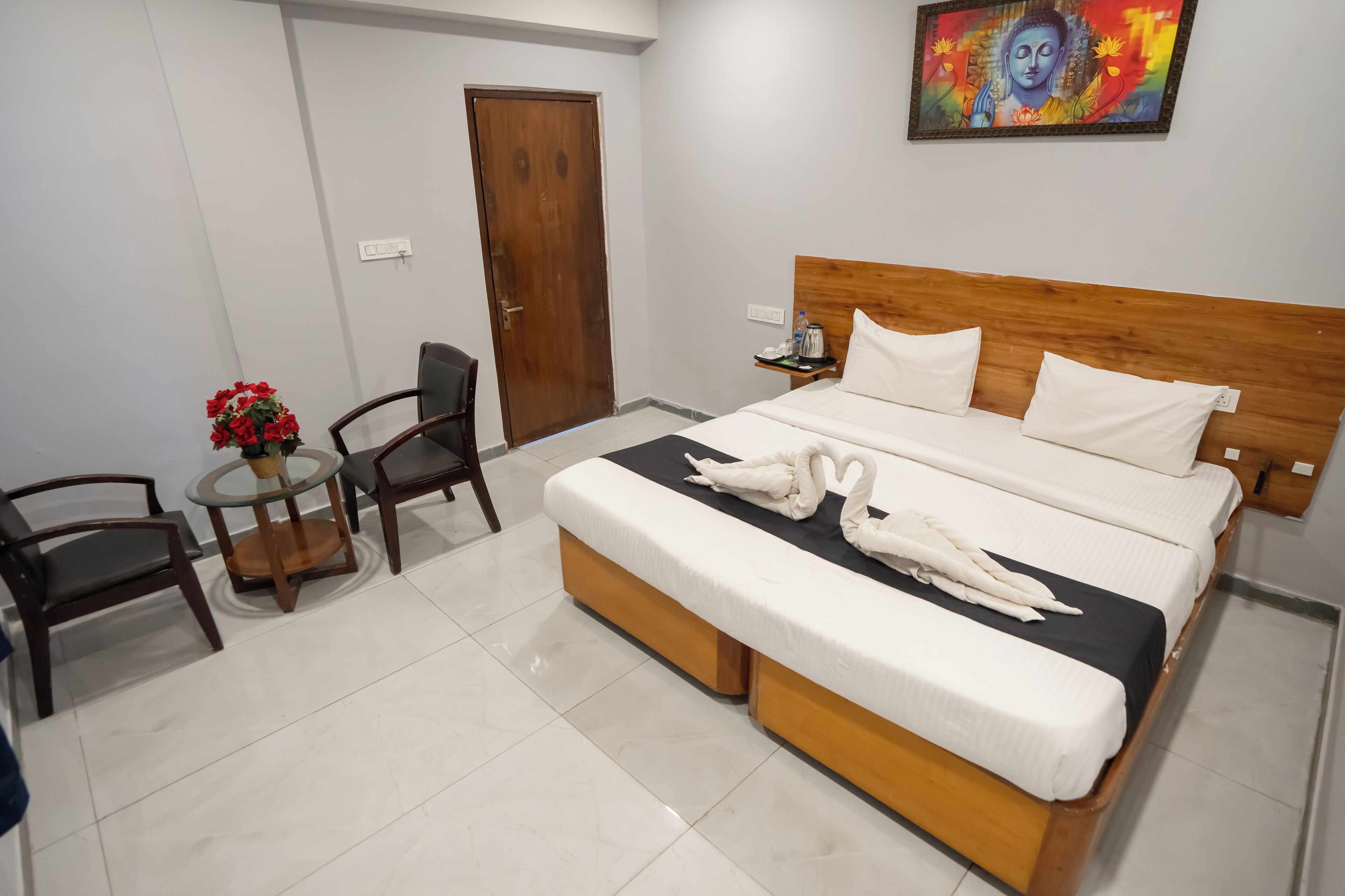 Habitación de hotel con una cama, dos sillas, una mesita con un florero y un cuadro en la pared.