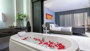 Une salle de bain moderne avec une grande baignoire remplie de pétales de roses, une douche en verre et un lit donnant sur le paysage urbain.
