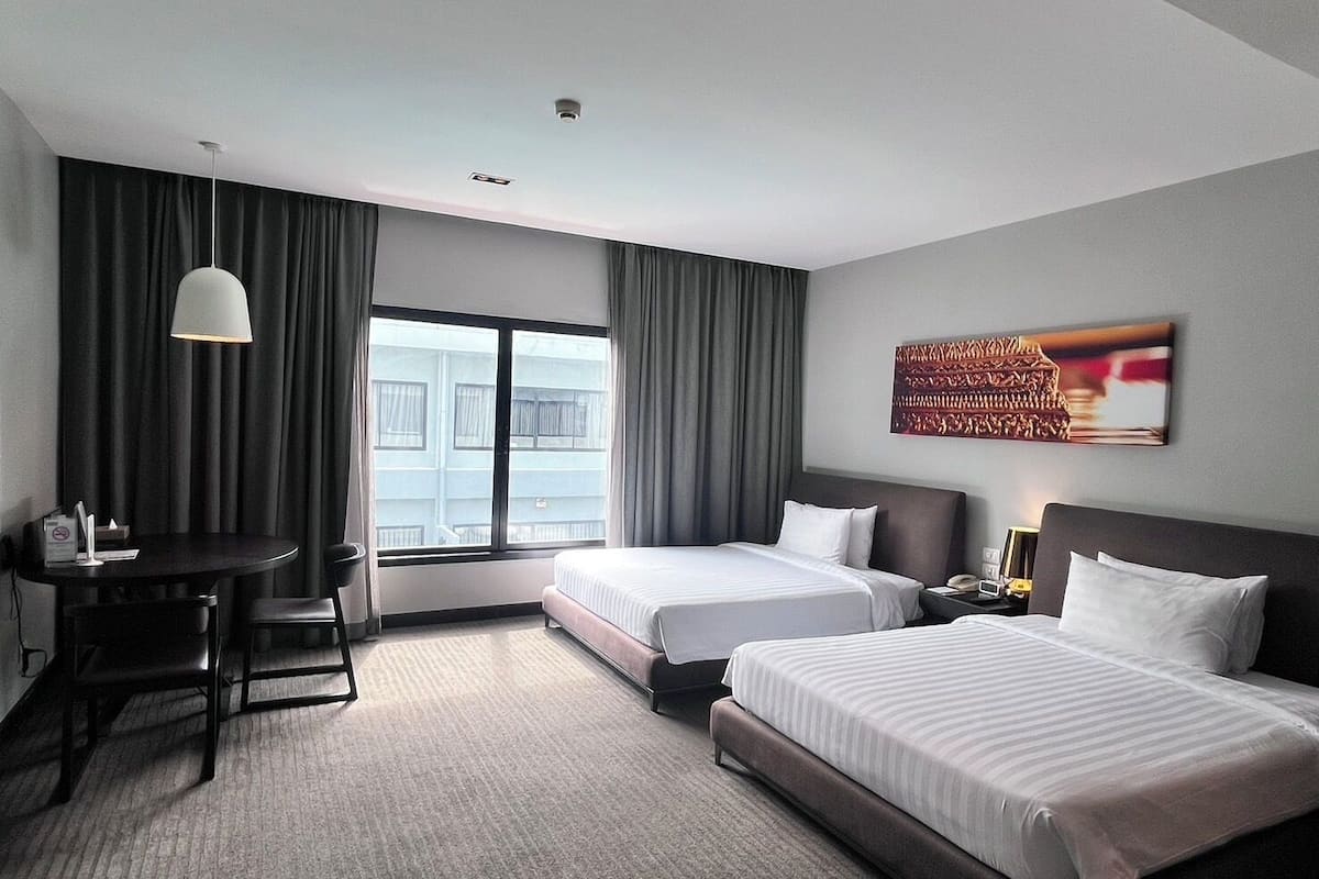 A hotel room with two beds, a desk, a chair, a lamp, and a window with curtains.