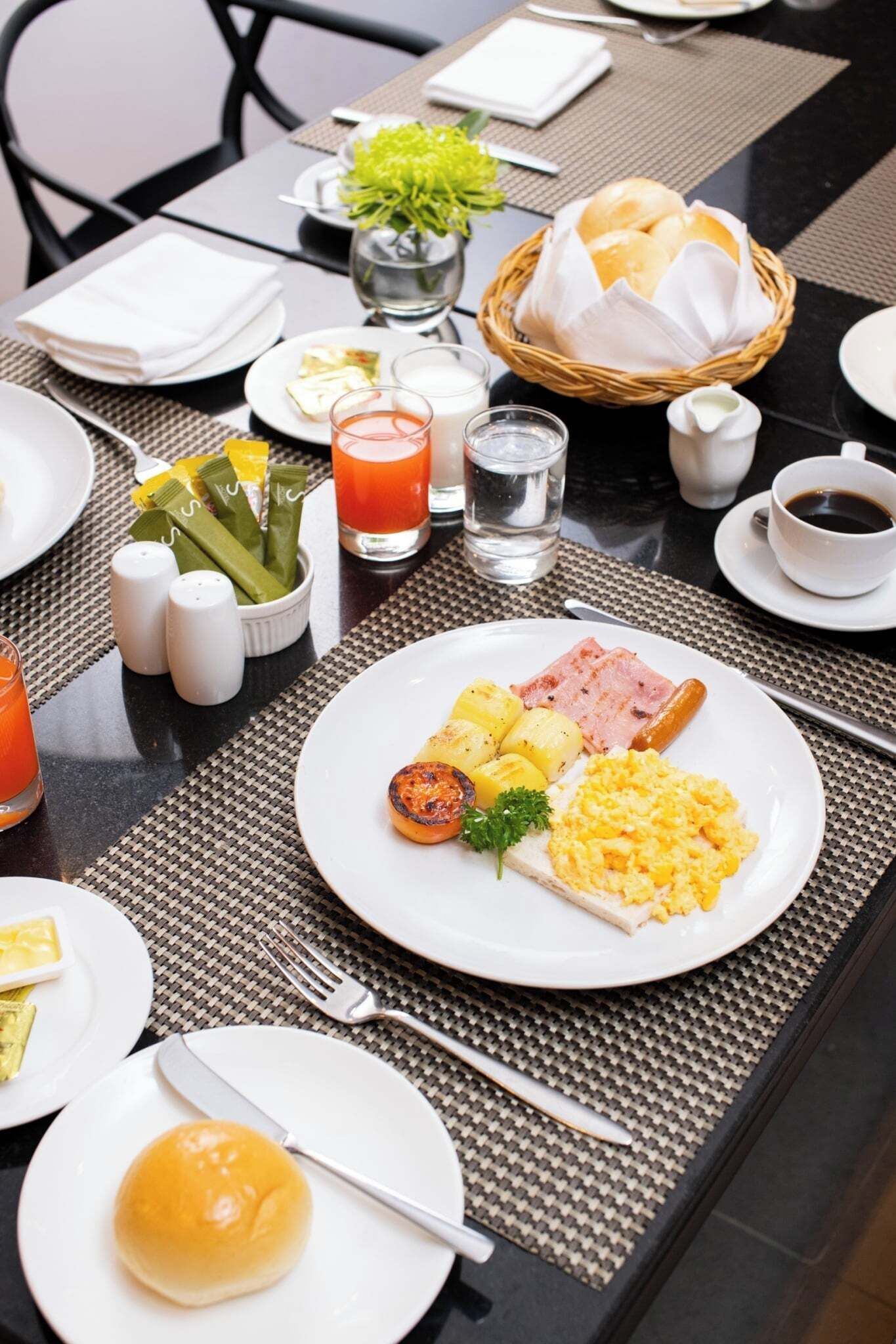 A breakfast setting with plates of eggs, ham, and potatoes, a basket of bread, and a glass of orange juice.