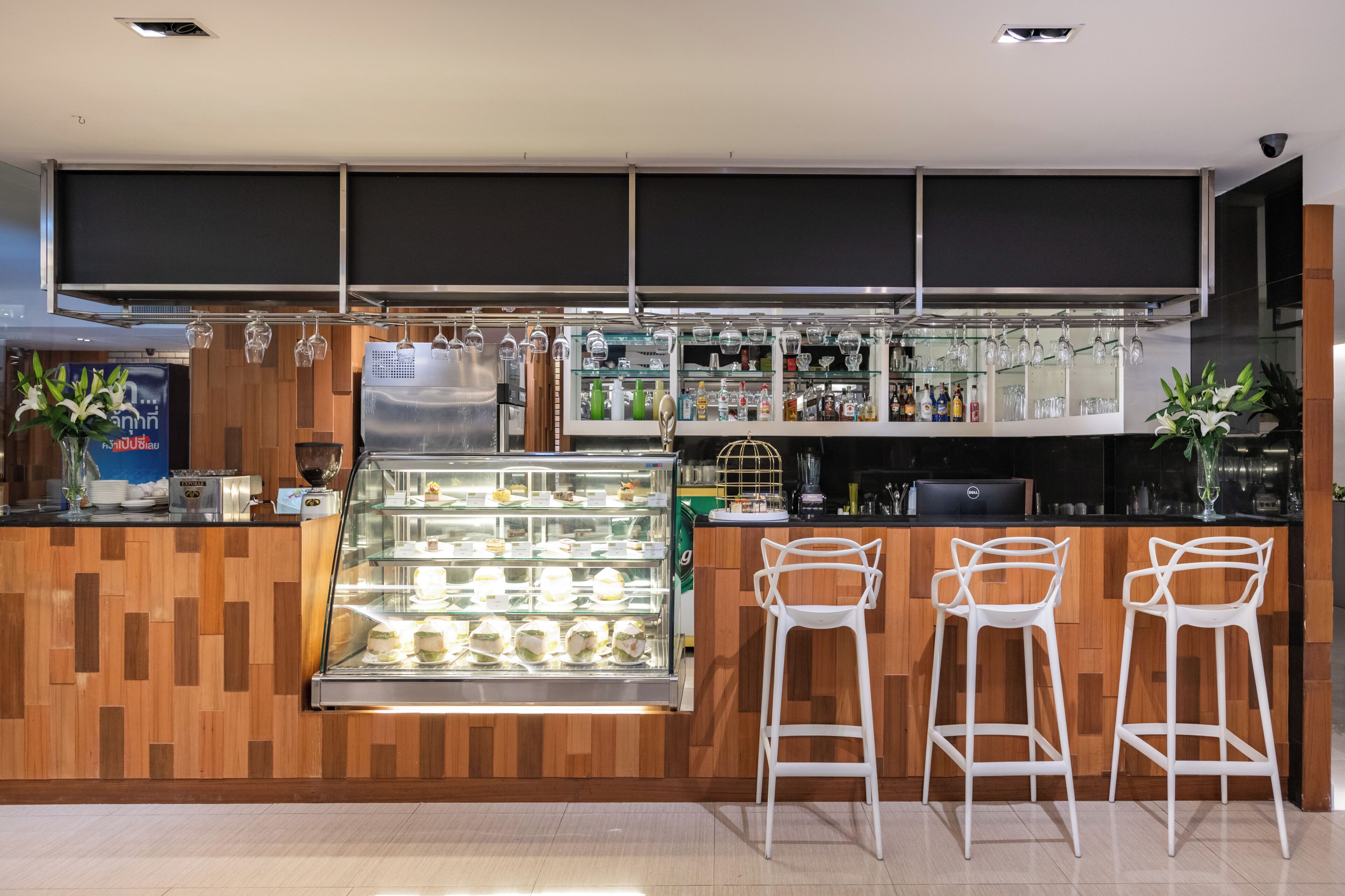 Una zona de bar moderna con una barra de madera, una vitrina de cristal con postres y taburetes blancos.