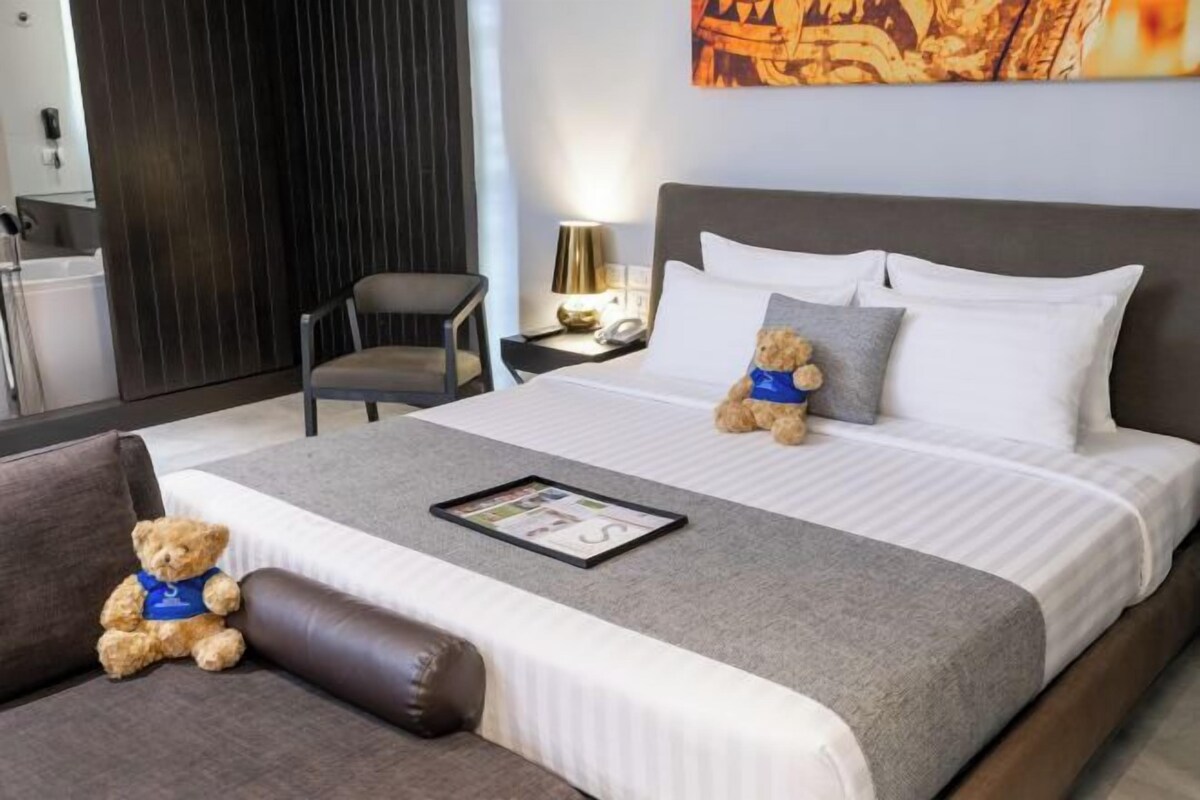 A neatly made bed with pillows, a bedside table with a lamp, a chair, and two teddy bears on the bed.