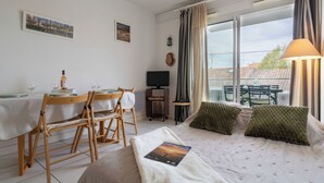 A cozy room with a bed, a dining table, and a TV. There are curtains and a window with a view of buildings. - Les Oyats by Interhome (Arcachon)