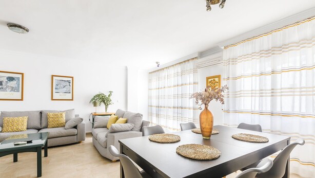A modern living room with a grey sofa, a coffee table, and a dining table with chairs. There are framed pictures on the wall and a vase with dried flowers on the dining table. - A&N Marisol (Torre del Mar)