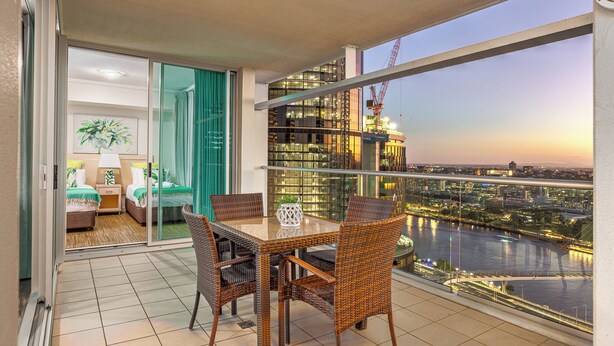 A balcony with a dining table and chairs, a sliding glass door leading to a bedroom, and a view of a cityscape and river at dusk.
