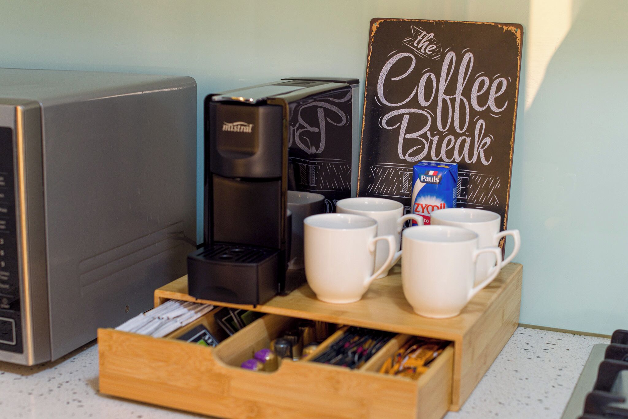 Een koffiehoek met een bord waarop 'The Coffee Break' staat, een koffiezetapparaat, verschillende witte mokken en een houten doos met pennen en potloden.