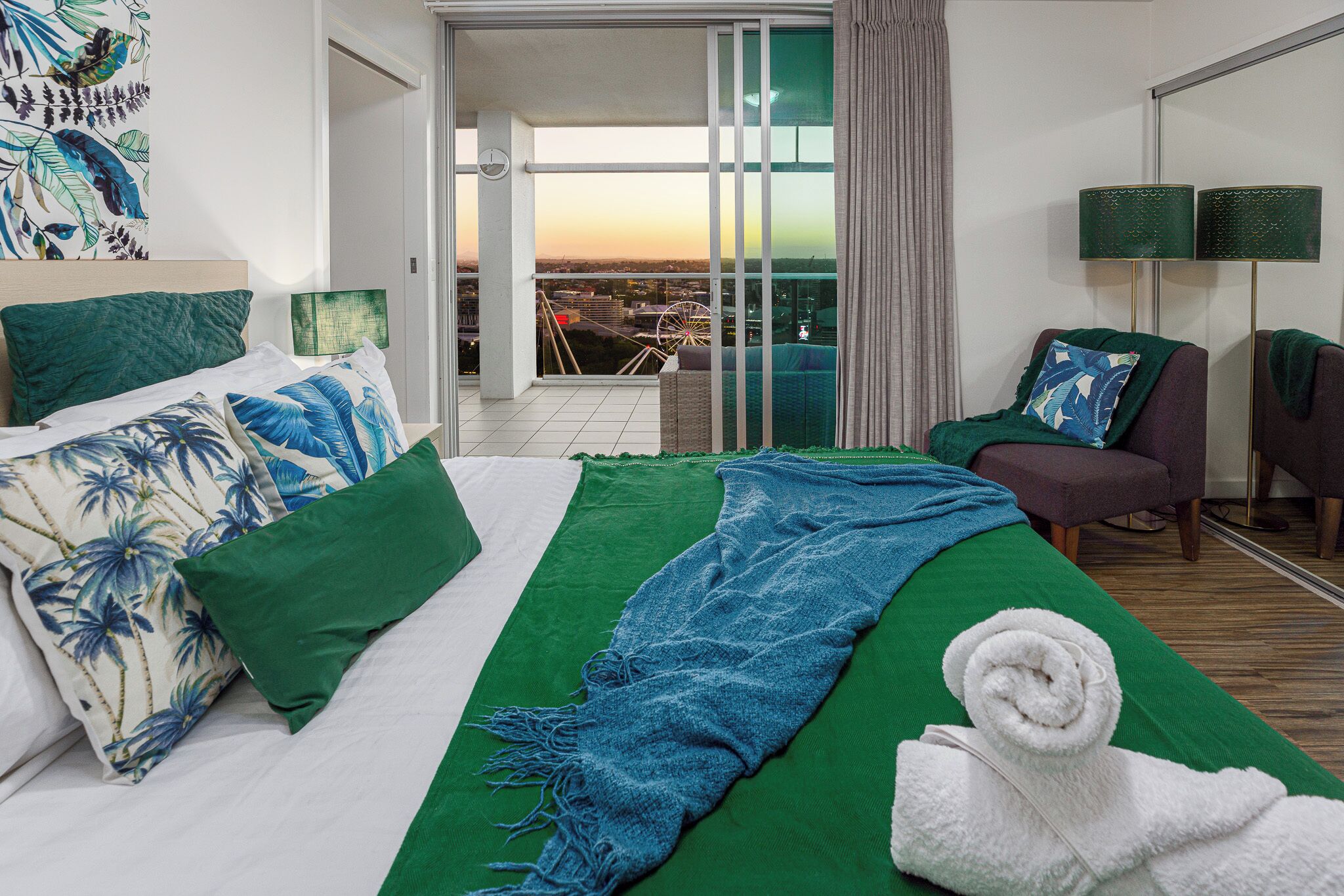 Habitación de hotel con cama, manta verde, toalla, silla y vista de una noria.