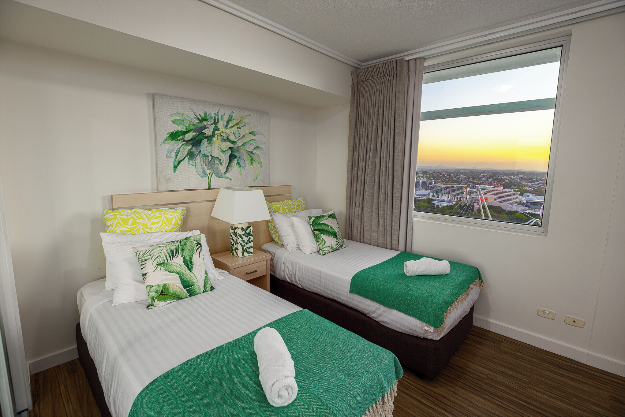 Habitación de hotel con dos camas, un amplio ventanal con vistas a la ciudad, un cuadro de una planta en la pared y una mesita de noche con lámpara.