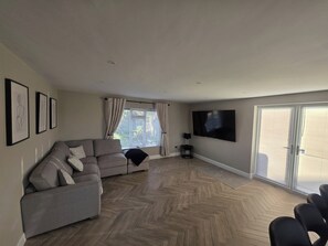 A living room with a grey sofa, a flat-screen TV mounted on the wall, and a herringbone-patterned wooden floor.