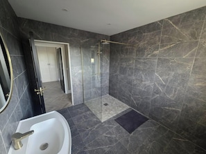 Un baño moderno con ducha de vidrio, lavamanos blanco y paredes de mármol oscuro.