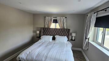 A bedroom with a large bed, a headboard, two bedside lamps, a window with curtains, and a herringbone-patterned floor.
