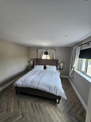 Un dormitorio con una cama grande, un cabecero, dos lámparas de noche, una ventana con cortinas y un suelo con estampado de espiga.