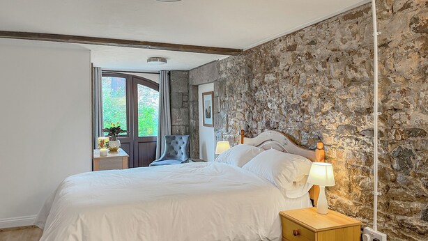 A bedroom with a stone wall, a bed with white linens, a wooden nightstand, and a chair.