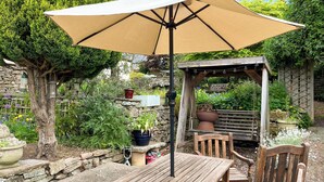 An outdoor patio with a wooden table and chairs, a beige umbrella, and a stone wall backdrop.