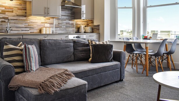Une cuisine moderne dotée d’une cuisinière intégrée, un coin repas avec une table et des chaises, et un canapé gris agrémenté d’un plaid.