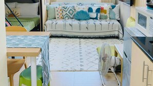 Un espace de vie compact comprenant un lit superposé, un canapé avec des coussins à motifs, une table à manger, un ventilateur et un four à micro-ondes.