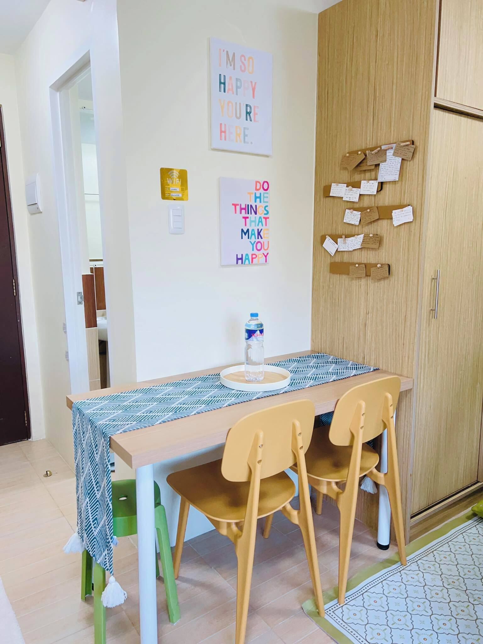 Een eetgedeelte met een houten tafel, vier stoelen en een tafelkleed met een patroon. Twee motiverende posters hangen aan de muur, een prikbord met notities en een waterfles op de tafel.