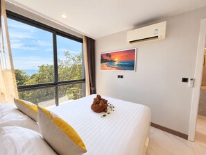 A hotel room with a large window, a painting of a sunset, an air conditioning unit, and a bed with pillows. - A303 - 1 Bedroom Serviced Apartment Partial Sea View at Ao Nang Beach (Tambon Ao Nang)