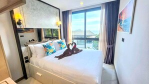 A hotel room with a bed, pillows, a nightstand, a lamp, a window with curtains, and a painting on the wall. - A303 - 1 Bedroom Serviced Apartment Partial Sea View at Ao Nang Beach (Tambon Ao Nang)