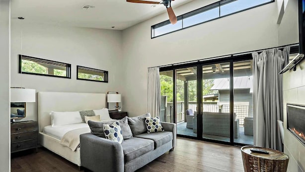 A modern bedroom with a bed, a grey sofa, a small table, and a large sliding glass door leading to a balcony.