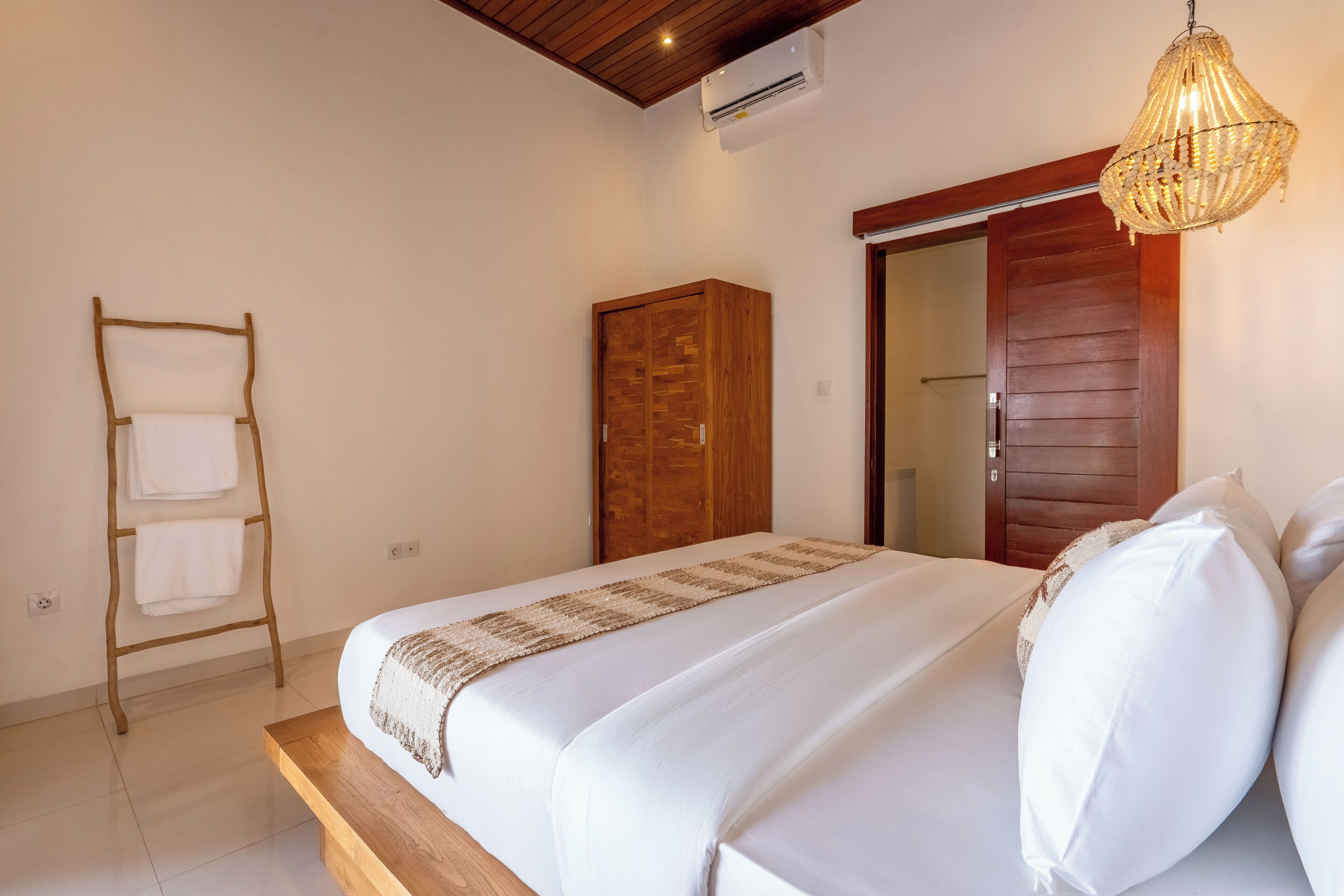 A neatly arranged bedroom with a wooden bed, a wooden wardrobe, a towel rack, and a hanging light fixture.