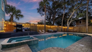 A swimming pool with a wooden deck, lounge chairs, and a fence with lights.