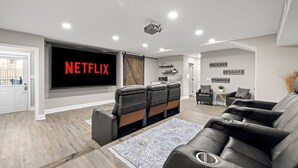 A modern living room with a large flat-screen TV displaying 'NETFLIX', a cozy seating area with leather sofas, a small coffee table, and a decorative rug. - 3BR Guest Escape w/ Pool & Theater (Palm Harbor)