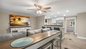 A modern kitchen with white cabinets, a dining area with a wooden table and gray seating, and a ceiling fan. - 3BR Guest Escape w/ Pool & Theater (Palm Harbor)