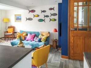 Un salon avec un canapé bleu, des coussins colorés, une chaise en bois, une petite table avec une lampe et un mur décoré d’œuvres d’art murales en forme de poisson.