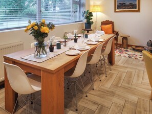 Un coin repas avec une table en bois dressée pour quatre personnes, entourée de chaises blanches, et une vue sur les arbres par la fenêtre.