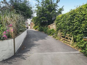 Une rue étroite bordée de maisons d’habitation, avec un mur blanc d’un côté et une clôture en bois de l’autre, menant à une maison dont on aperçoit le portail du garage.