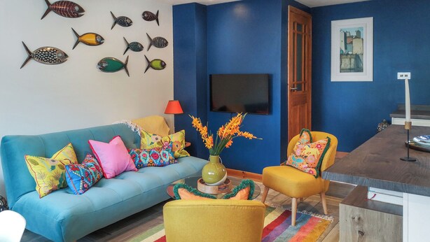 A living room with a blue sofa, colorful cushions, a yellow armchair, a wooden coffee table with a vase of flowers, and a television mounted on the wall. - 2 bedroom accommodation in Barnstaple (Barnstaple)