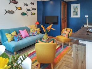 Un salon avec un canapé bleu, des coussins colorés, un fauteuil jaune, une table basse en bois ornée d’un vase de fleurs et un téléviseur fixé au mur.