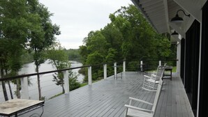 Una terraza de madera con una mesa y mecedoras, con vista a un río y rodeada de árboles.