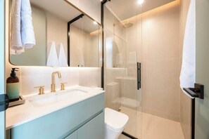 Un baño moderno con cabina de ducha de vidrio, lavamanos blanco con detalles dorados y un espejo grande.