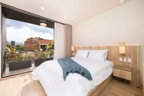 Una habitación de hotel moderna con una cama grande, cabecera de madera, mesita de noche y vistas a la ciudad a través de ventanales.