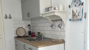 Et lille køkken med lyse skabe, en træbordplade og en indbygget vask. Kopperviser og en lille hylde over vasken.