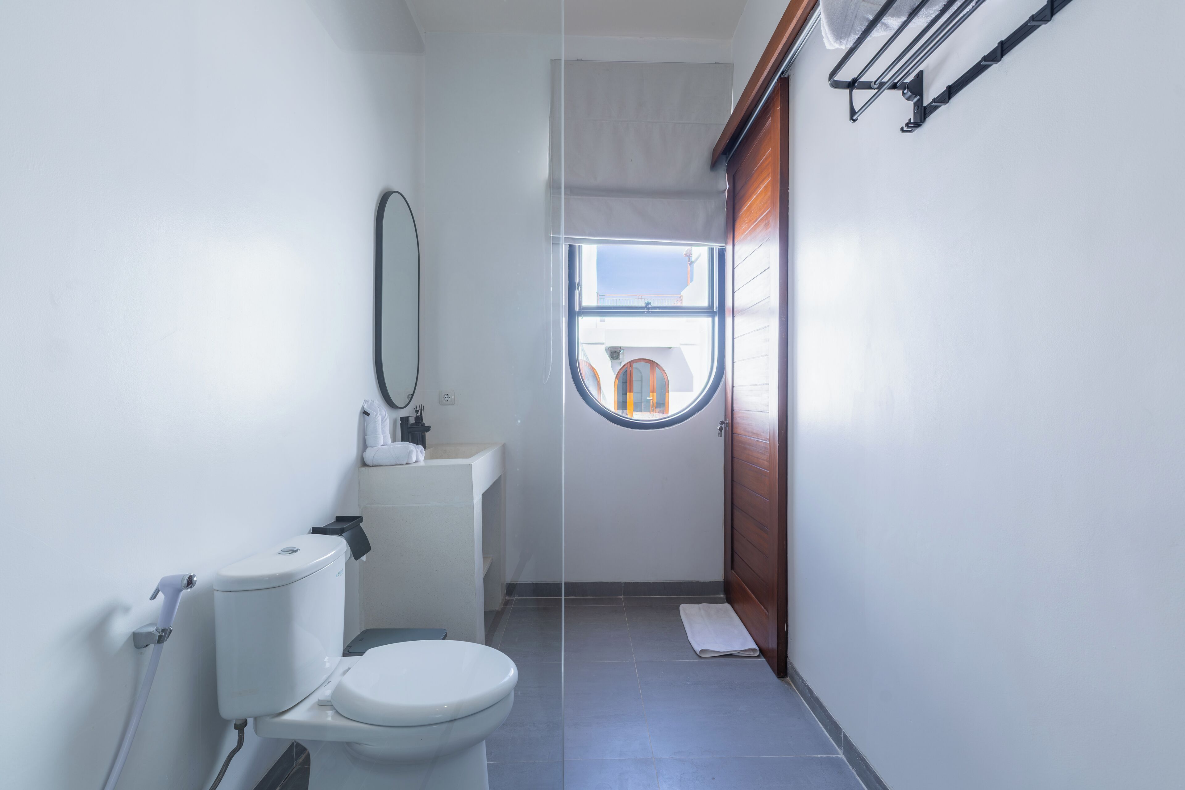 Une petite salle de bain comprenant des toilettes, un lavabo et un miroir. Une fenêtre laisse entrer la lumière naturelle et offre une vue sur l’extérieur.