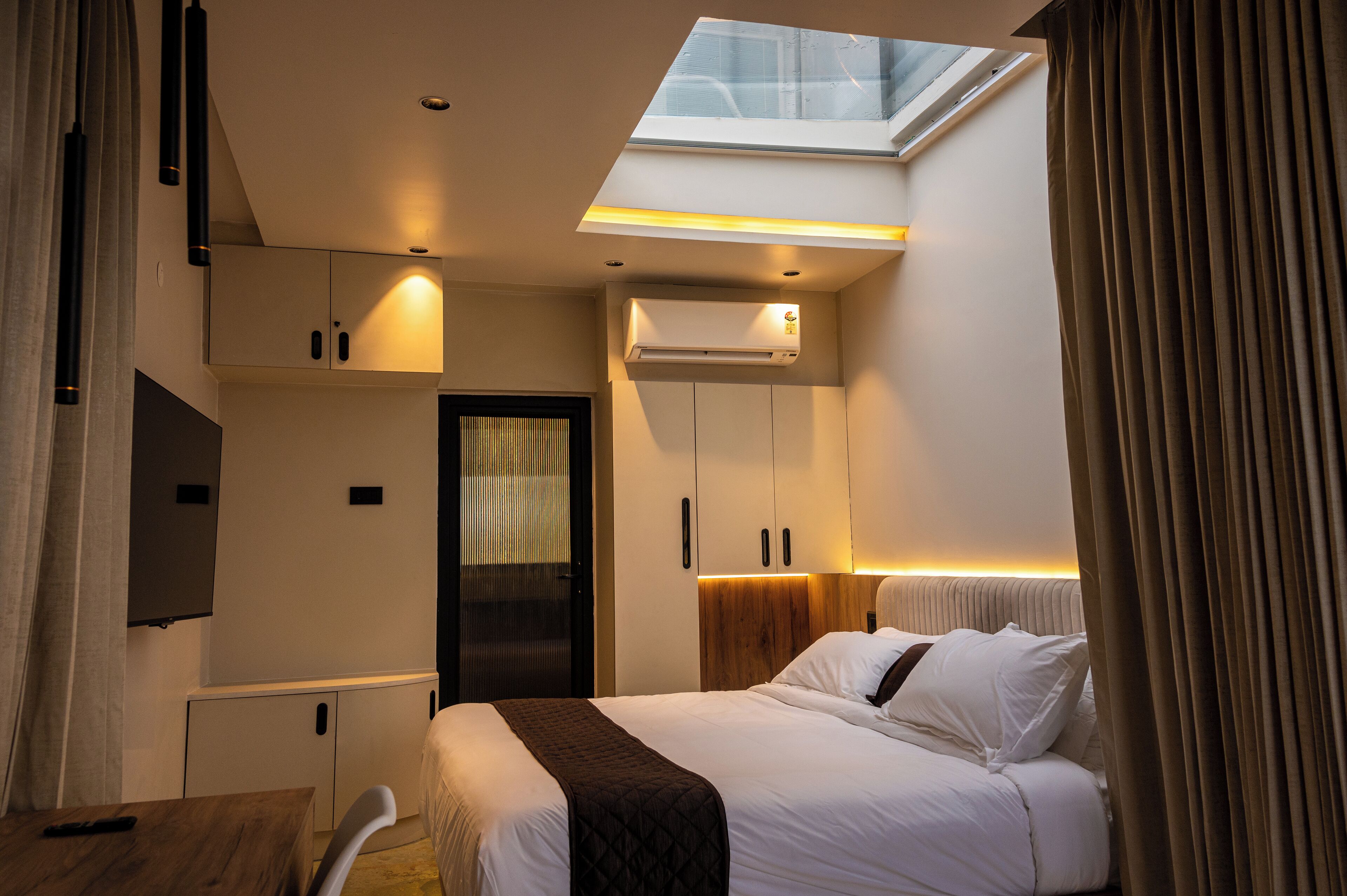 A hotel room with a bed, a desk, a TV, an air conditioning unit, and a skylight.