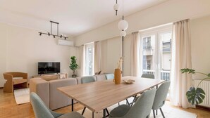 A modern living room with a wooden dining table, grey upholstered chairs, a sofa set, a TV, and a potted plant. - Central Downtown Living (Athina)