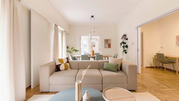 A modern living room with a beige sectional sofa, a round coffee table, and a dining area in the background. - Central Downtown Living (Athina)