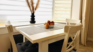 A dining area with a white table, two chairs, a bowl of fruit, and a vase with dried plants.